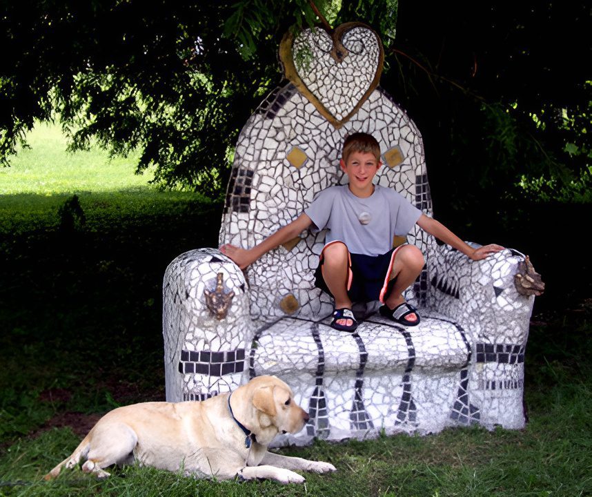 Boy sitting on mosaic chair, dog nearby.